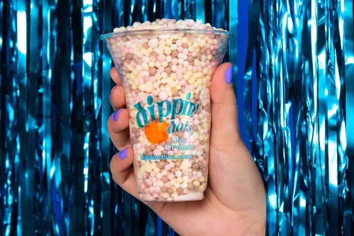 A cup of colorful Dippin' Dots held by a hand in front of blue streamers.
